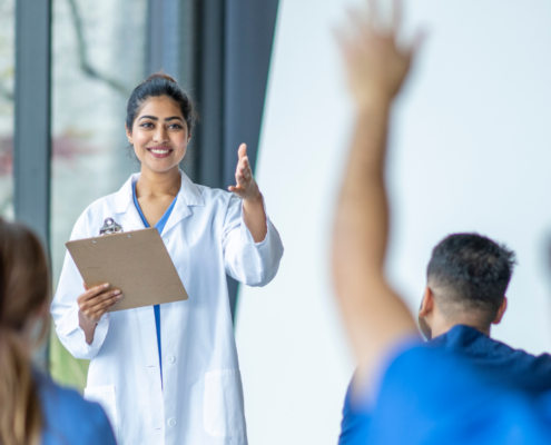 Uma médica mulher com uma prancheta em sua mão esquerda com o braço direito estendido em direção a um aluno de medicina com o braço estendido para fazer uma dúvida. O ambiente retrata uma experiência acadêmica