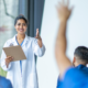 Uma médica mulher com uma prancheta em sua mão esquerda com o braço direito estendido em direção a um aluno de medicina com o braço estendido para fazer uma dúvida. O ambiente retrata uma experiência acadêmica