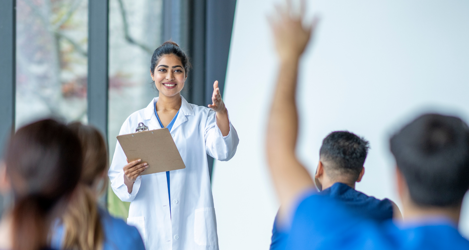 Especialização InCor - Experiência Acadêmica Uma médica mulher com uma prancheta em sua mão esquerda com o braço direito estendido em direção a um aluno de medicina com o braço estendido para fazer uma dúvida. O ambiente retrata uma experiência acadêmica
