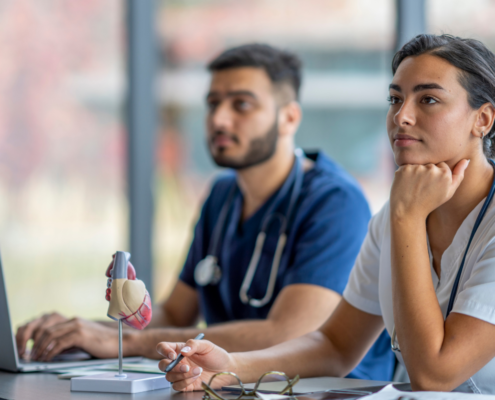 Dois estudantes, um homem jovem branco com barba e uniforme de médico azul escuro ao fundo da imagem; uma mulher jovem de pele bronzeada e cabelos castanhos lisos e presos no plano da frente estão em um ambiente de sala de aula para representar a especialização com bolsa no Ensino InCor para multiprofissionais