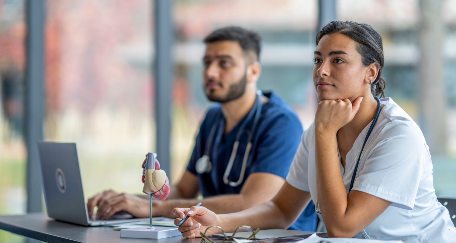 Especialização com Bolsa no InCor - Ensino InCor Dois estudantes, um homem jovem branco com barba e uniforme de médico azul escuro ao fundo da imagem; uma mulher jovem de pele bronzeada e cabelos castanhos lisos e presos no plano da frente estão em um ambiente de sala de aula para representar a especialização com bolsa no Ensino InCor para multiprofissionais
