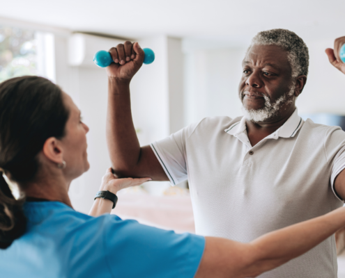 Uma fisioterapeuta está na esquerda da imagem vestindo um jaleco branco enquanto realiza uma fisioterapia cardiorrespiratória num paciente homem negro que veste uma polo branca com cabelos grisalhos com os braços levantados e semi dobrados com dois halteres pequenos em cada mão.