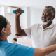 Uma fisioterapeuta está na esquerda da imagem vestindo um jaleco branco enquanto realiza uma fisioterapia cardiorrespiratória num paciente homem negro que veste uma polo branca com cabelos grisalhos com os braços levantados e semi dobrados com dois halteres pequenos em cada mão.