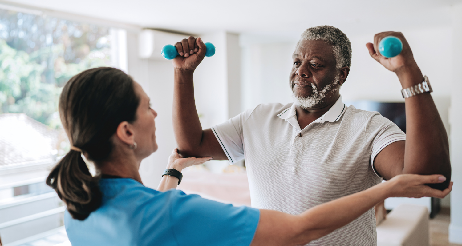 Uma fisioterapeuta está na esquerda da imagem vestindo um jaleco branco enquanto realiza uma fisioterapia cardiorrespiratória num paciente homem negro que veste uma polo branca com cabelos grisalhos com os braços levantados e semi dobrados com dois halteres pequenos em cada mão.