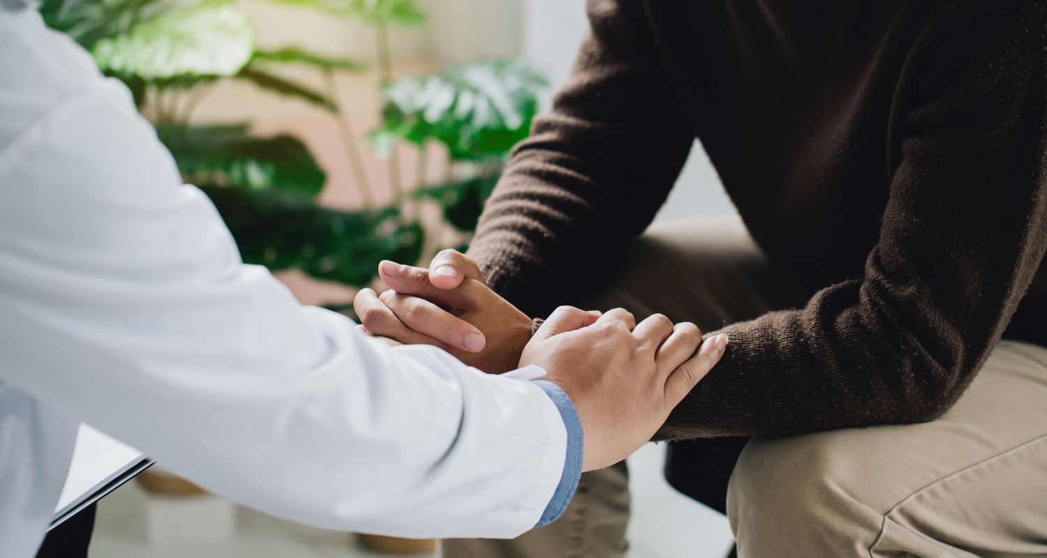 Psicologia Hospitalar Ensino InCor Imagem com aproximação onde mostra apenas os braços e as pernas do médico e do seu paciente sentado em sua frente. O médico, que retrata o papel de um profissional de psicologia hospitalar, está a esquerda da imagem com as mãos colocadas sob os antebraços do paciente que está na direita da imagem. O médico usa jaleco branco e tem a pele clara enquanto o paciente tem a pele mais escura e usa uma blusa de manga comprida marrom escuro. Eles tão num consultório claro que tem plantas ao fundo.