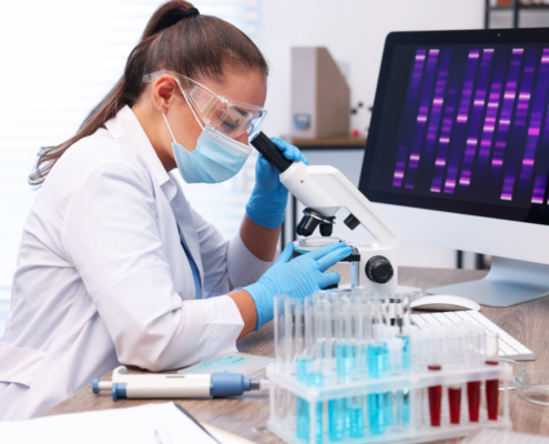 Consultório médico onde uma médica com pele clara, cabelos lisos em castanho escuro amarrados vestindo um jaleco branco, mascara e luvas em tom de azul claro, está realizando uma análise de teste genético um instrumento microscópico
