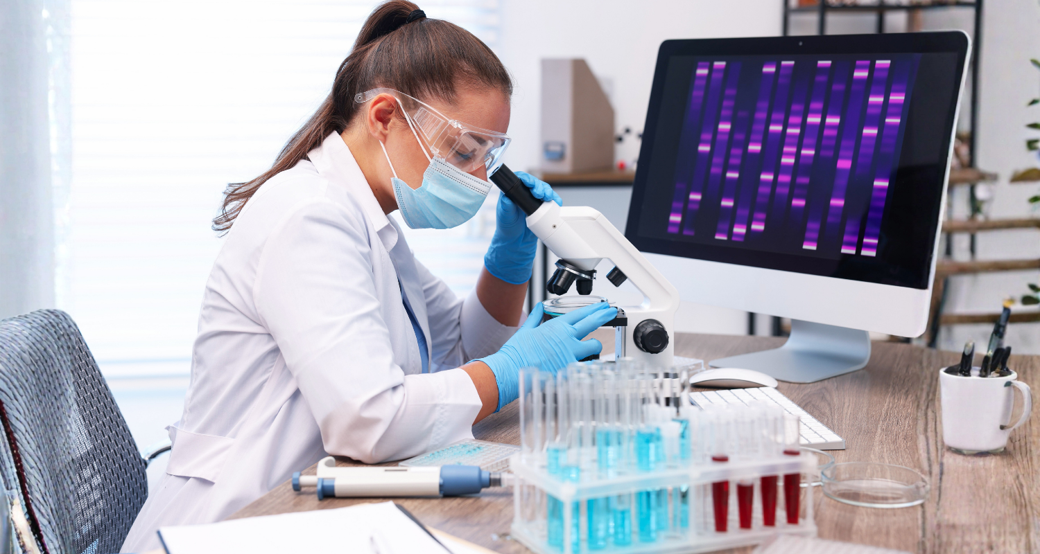 Consultório médico onde uma médica com pele clara, cabelos lisos em castanho escuro amarrados vestindo um jaleco branco, mascara e luvas em tom de azul claro, está realizando uma análise de teste genético um instrumento microscópico