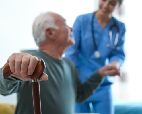 Uma médica ao fundo dando suporte a um paciente idoso homem que está no plano na frente e em destaque para enfatizar o cuidado de cardiogeriatria