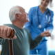 Uma médica ao fundo dando suporte a um paciente idoso homem que está no plano na frente e em destaque para enfatizar o cuidado de cardiogeriatria