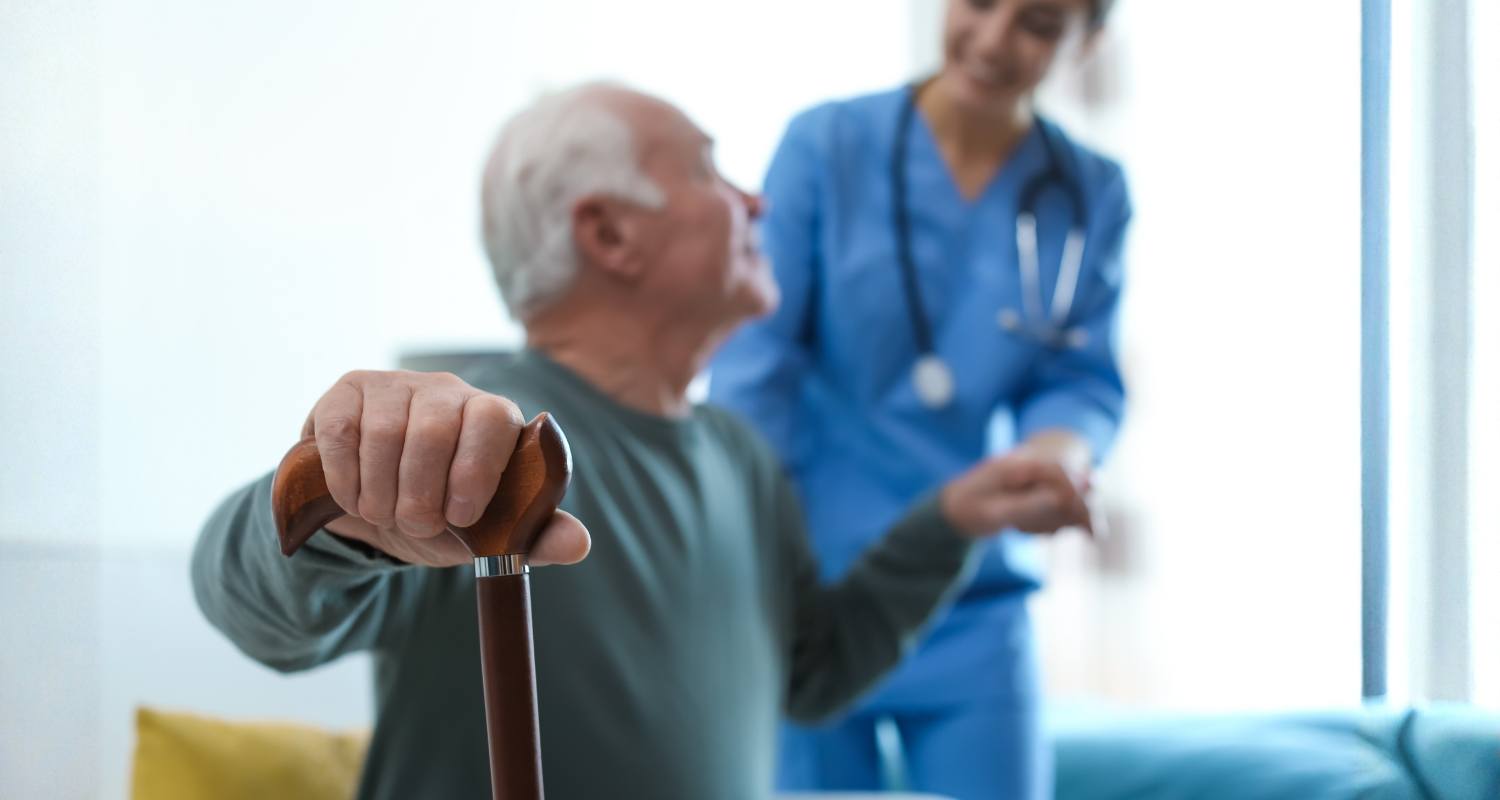 Uma médica ao fundo dando suporte a um paciente idoso homem que está no plano na frente e em destaque para enfatizar o cuidado de cardiogeriatria