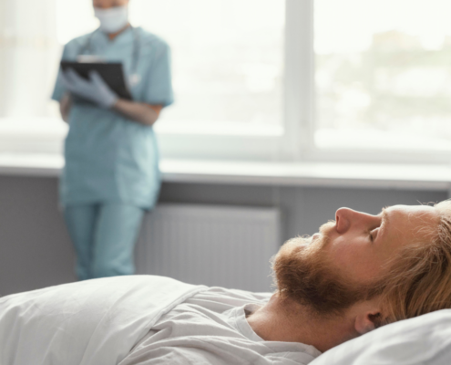 Paciente homem adulto de pele clara e cabelos louros dormindo no leito hospitalar. Ao fundo, está uma médica fazendo anotações representando a área de medicina do sono