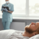 Paciente homem adulto de pele clara e cabelos louros dormindo no leito hospitalar. Ao fundo, está uma médica fazendo anotações representando a área de medicina do sono