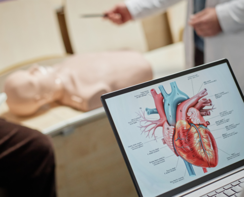 Imagem fechada de uma aula na especialização em cardiologia. No canto direito temos uma tela de notebook com a imagem de um coração e a descrição de sua anatomia. Ao fundo, um pouco desfocado, temos a imagem de um boneco para sessões práticas com um professor apontado para ele